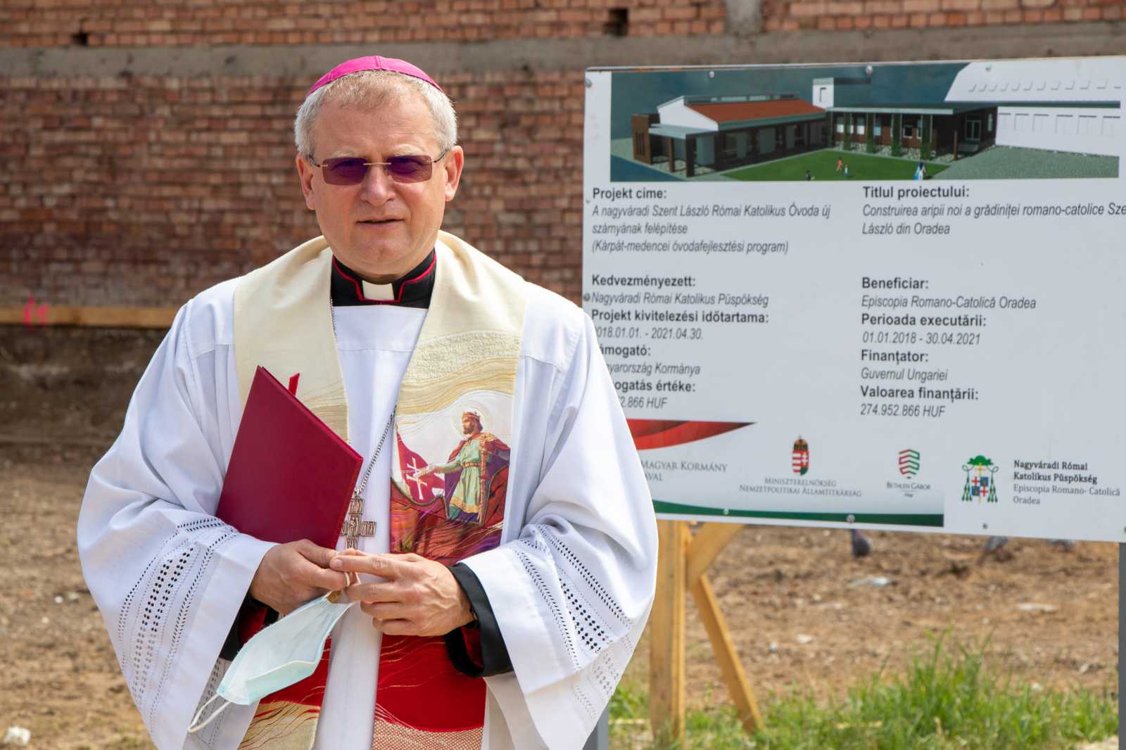 A püspök immár az építkezés helyszínén, a beruházást jelző pannó előtt