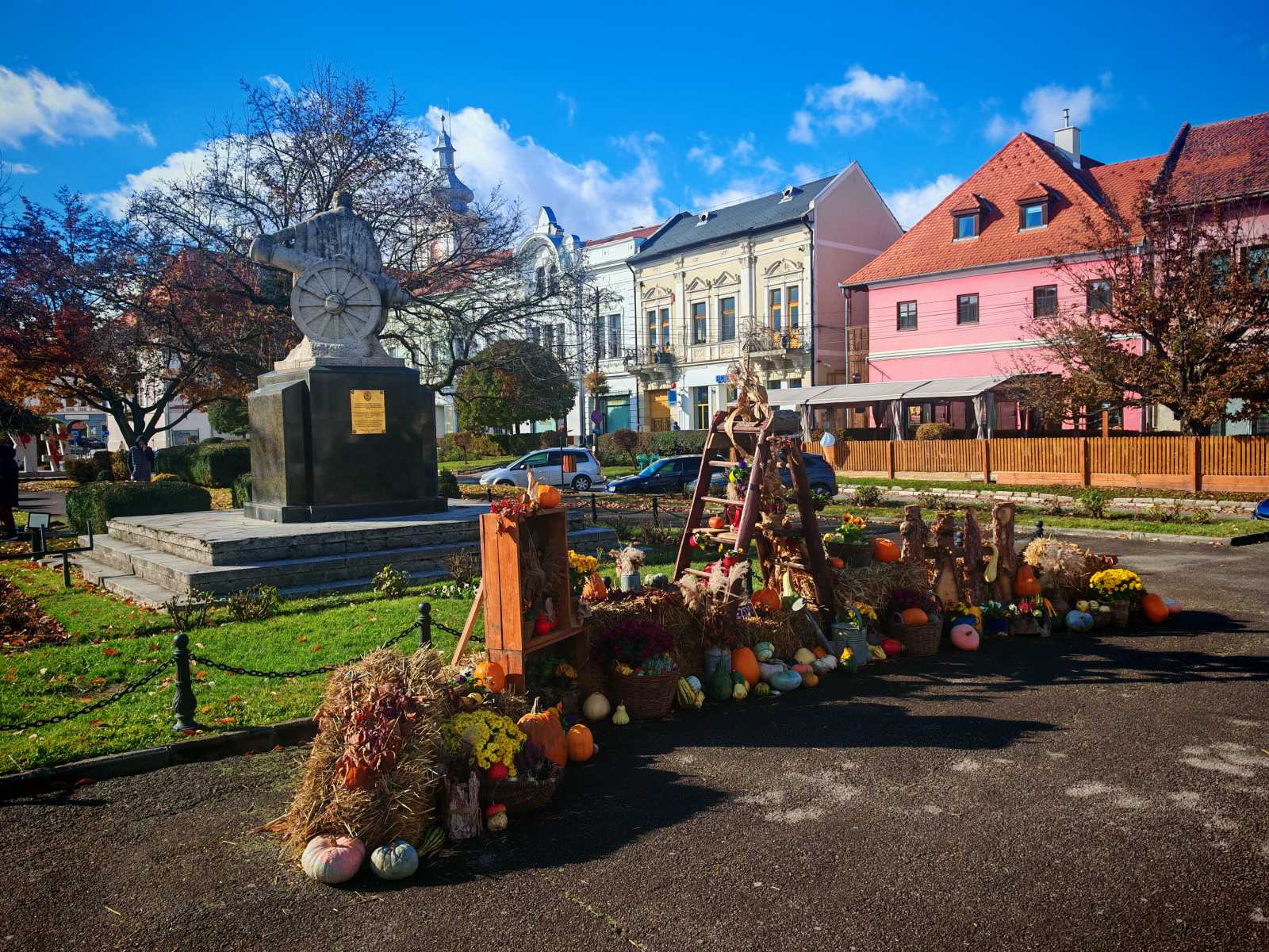A fotó Kézdivásárhely főterén készült, ahol a történelmi városközpont impozáns épületei között egy hangulatos őszi installáció kapott helyet. A szobor előtti térre szalmabálák, tökök, kukoricák, krizantémok és más őszi termények kerültek, a díszítés pedig a falusi gazdálkodás és a természet bőségének szimbóluma. A napfényben ragyogó színek és a gondosan elrendezett dekorációk előtt a járókelők szívesen időznek és fényképezkednek.